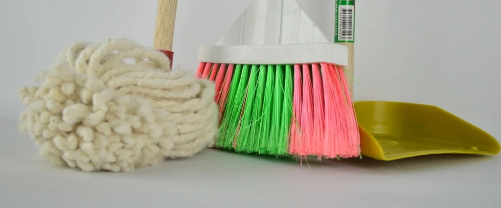 A set of cleaning tools including a white mop, a colorful broom with pink and green bristles, and a yellow dustpan, standing against a light background.