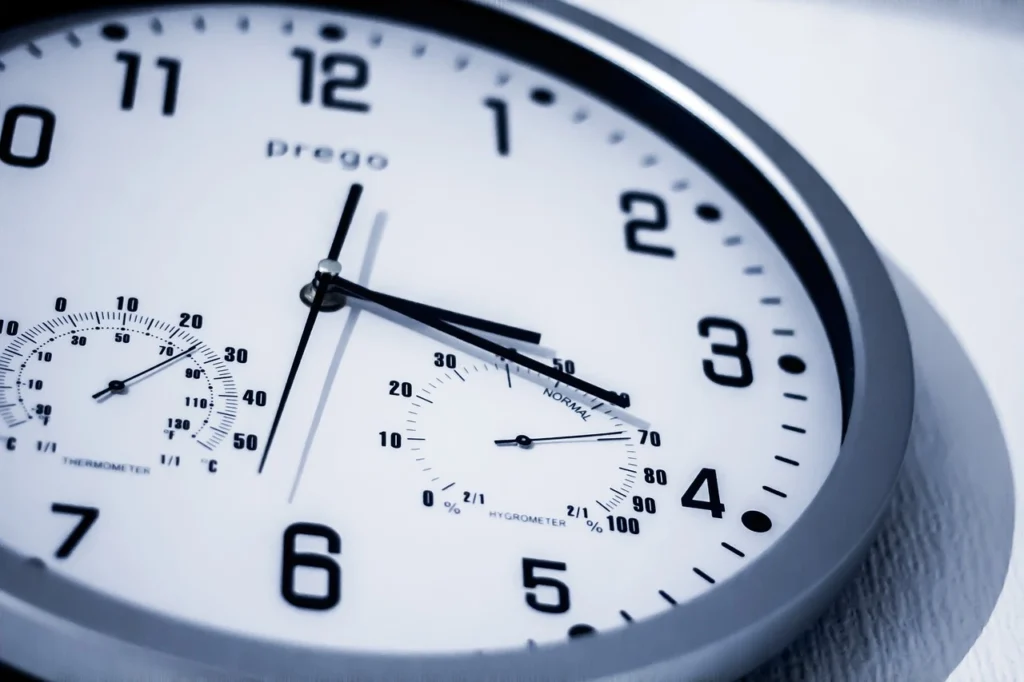 Close-up view of a round wall clock displaying both time and temperature. The clock features black hour, minute, and second hands on a white face, with bold black numbers and small dials showing humidity and temperature readings