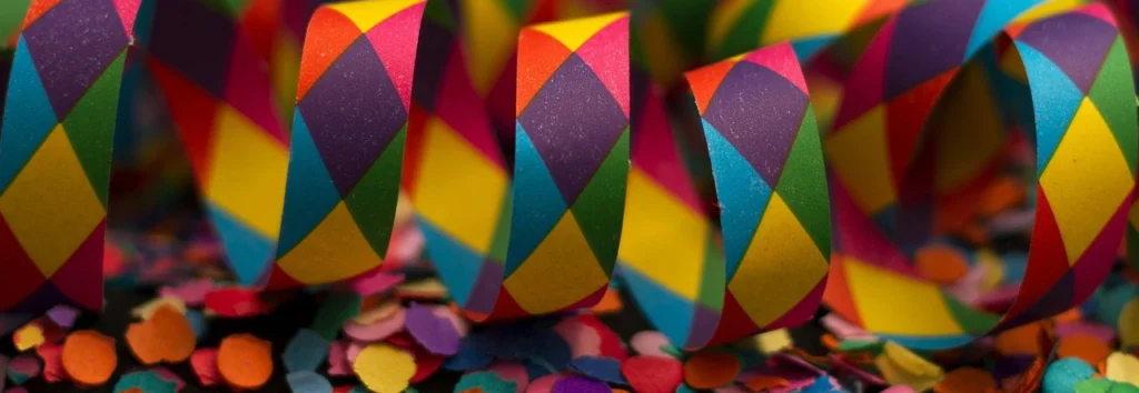 Vibrant multicolored streamers featuring a geometric diamond pattern in red, green, blue, purple, and yellow, lying among scattered colorful confetti.