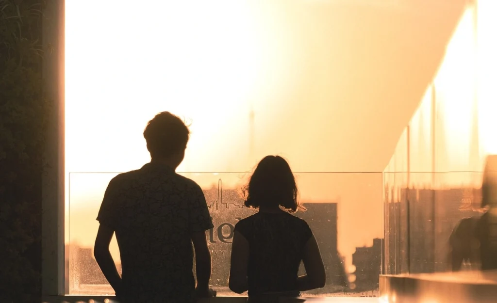 A couple stands together by a window in their well-maintained apartment, watching a beautiful sunset illuminate the horizon, reflecting their tenant satisfaction with the property.